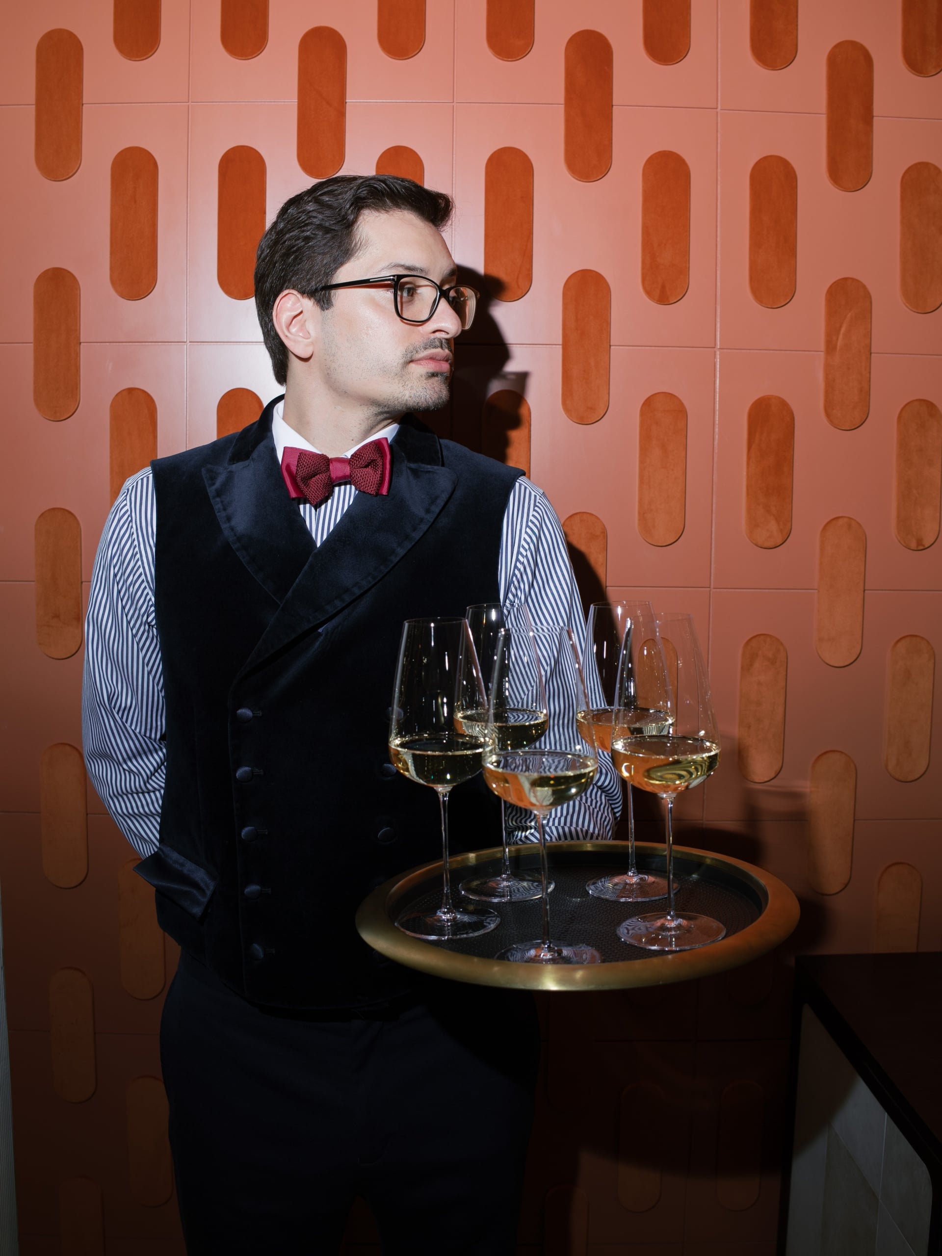 une personne en costume tenant un plateau avec des verres à vin, devant un fond rouge